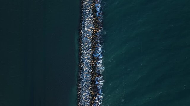 Aerial view of stone breakwater with waves hitting rocks in deep blue ocean water