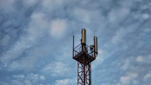 A red telecommunications tower with multiple antennas stands stationary against a dynamic blue sky. Slowly drifting clouds shift in shape and density above the structure throughout the scene.