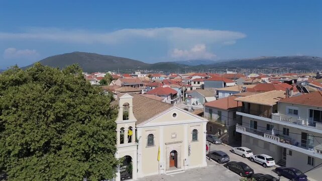 High aerial drone view of Lefkada town showing Evangelistria Orthodox church surrounded by traditional houses with red roofs and scenic mountains under clear blue summer sky.