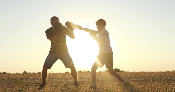 Silhouette father and son practice boxing and sparring together in rural field at sunset. They train outdoors, build confidence, enjoy outdoor exercise. Warm backlight creates energetic family fitness