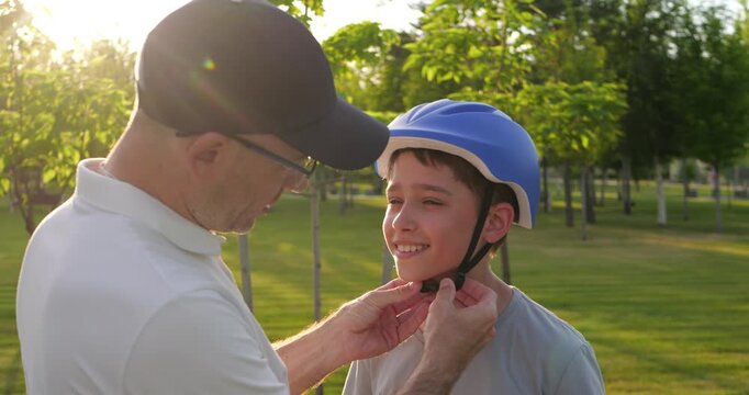 Father adjusts son's bicycle helmet in sunny park. Boy prepares for safe cycling with caring parent. Family shares joyful outdoor activity together in summer sunshine and active lifestyle.