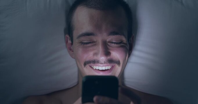 Top Down Portrait of Happy Young Caucasian Man Lying in Bed at Night Using Smartphone and Smiling