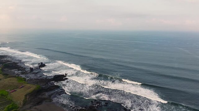 Drone footage of powerful ocean waves rolling and crashing onto the rocky black sand coastline of Kedungu Beach, Tabanan, Bali, Indonesia. Wide seascape with multiple wave lines