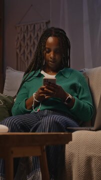 Vertical shot of young adult African American woman sitting on sofa at night, multitasking with smartphone and laptop. Clip is perfect for ads about digital burnout or remote work lifestyle