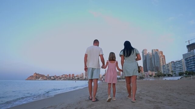 A family enjoys a walk along the beach at sunset in Altea. They hold hands while the waves gently touch the shore. The town and buildings are in the background.
