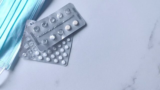 Woman's hand places a blister pack of pills on a table beside another blister pack and a medical face mask, healthcare concept.