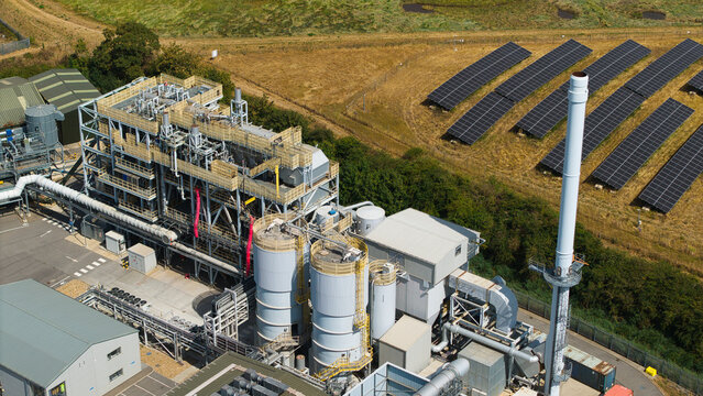 Aerial view of a biomass energy power plant featuring industrial structures, pipes, and a tall chimney, situated next to a field of solar panels in Boston, England, United Kingdom.