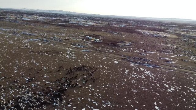 Aerial view sweeping over vast tundra landscape with scattered snow patches, dirt roads, and distant mountains under overcast sky in Mini Gobi, Mongolia