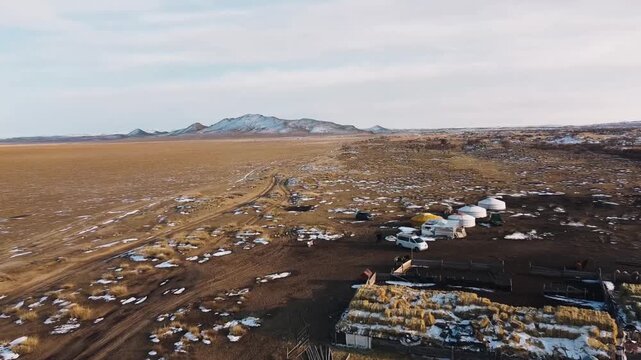 Aerial view of yurts next to dirt road cutting through vast steppe grassland with patches of snow and distant mountains under overcast sky in Mini Gobi, Mongolia