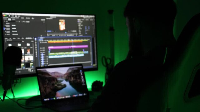 Exhausted video editor pauses work in dark green-lit studio, head in hands at desk with monitor, timeline and laptop. Concept of burnout, stress, deadlines, late-night editing.
