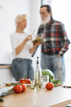 An older couple in casual clothing is in a bright kitchen, preparing a fresh salad with various vegetables and ingredients on a wooden countertop