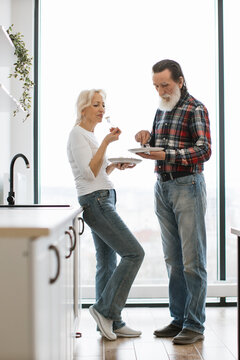 A senior man and woman stand in a bright kitchen, sharing a meal and enjoying each other's company