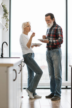 An older couple enjoys a meal together in their modern kitchen, standing by the counter near a large window