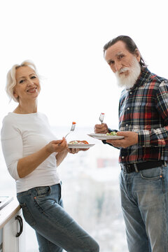 A happy senior couple stands near a window, each holding a plate of food and a fork, sharing a moment together