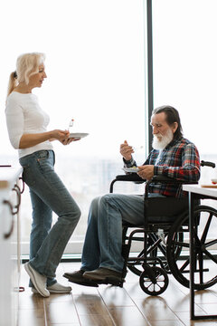 An older couple shares a meal by a large window, one in a wheelchair, highlighting companionship and care