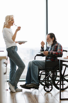 An elderly couple enjoys a meal together, with the man seated in a wheelchair and the woman standing nearby