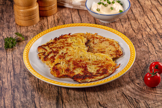 Crispy golden potato pancakes draniki served on white plate with yellow rim, sour cream, cherry tomatoes, and parsley on rustic wooden table