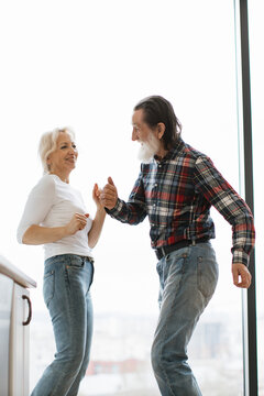 An energetic elderly couple shares a joyful moment dancing and laughing together indoors near a large window