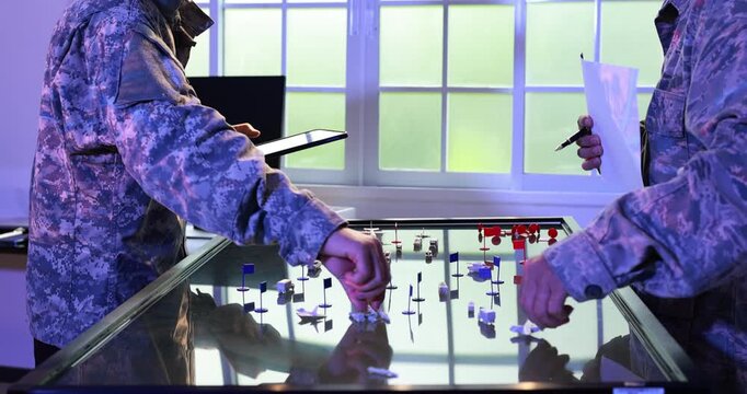 Two people in camouflage uniforms play a tabletop strategy game with miniature pieces in a brightly lit room, suggesting military training and tactical planning.