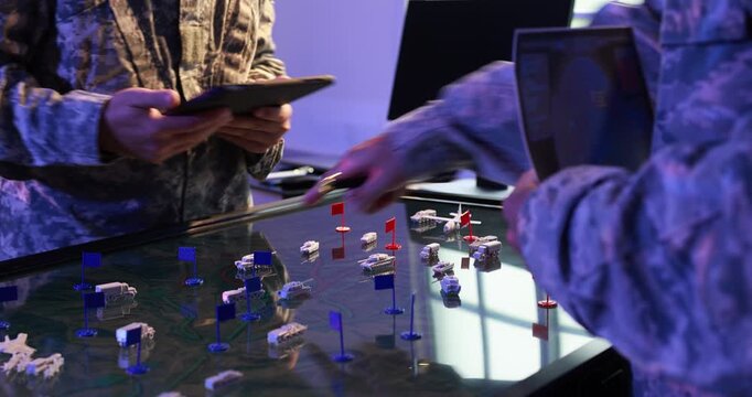 Military personnel use a digital map table with unit markers while one holds a tablet during a tactical planning meeting.