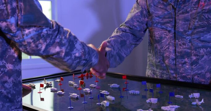 Two uniformed military personnel plan an operation on an illuminated digital terrain table with miniature vehicles, buildings and colored tactical markers.