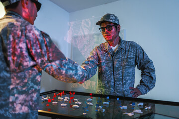 Naklejka premium Two uniformed soldiers review a digital tactical map table, planning a military operation in a modern command room.