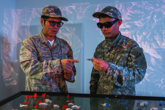 Two soldiers in camouflage uniforms and dark glasses study a tabletop military map during a tactical planning exercise under blue and red light.