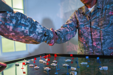 Naklejka premium Two soldiers in camouflage shake hands over a military strategy table with miniature vehicles and red and blue flags, planning, teamwork and agreement.