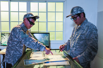 Two military personnel in camouflage uniforms review a large map on a lighted planning table in an...