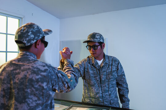 Two uniformed soldiers wearing sunglasses greet each other with a fist bump in a bright indoor room beside a window and table.