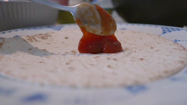 Person spreading tomato sauce on a round tortilla base using a spoon for homemade pizza preparation.