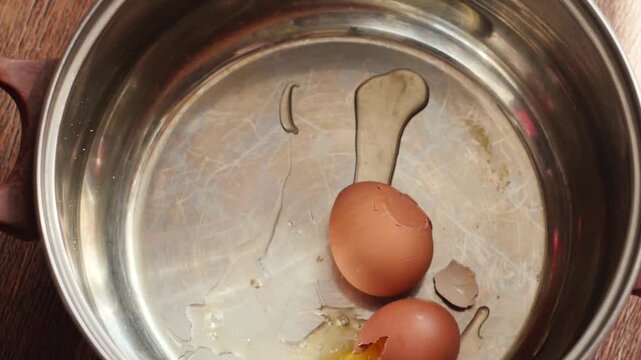 A second raw brown chicken egg falls into a metal cooking pot from above alongside a previously broken egg. Upon impact, the shell cracks