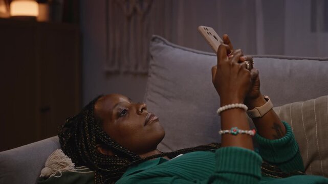 African American young adult woman lying on sofa and scrolling on smartphone at night. Footage represents digital burnout, social media addiction, or remote work fatigue for mental health campaigns