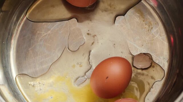 A third raw brown chicken egg falls into a metal pot containing broken shells and spilled liquid egg white. The egg impacts the bottom of the pan, adding to the messy culinary preparation process.