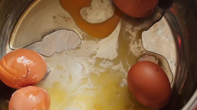 Another raw brown chicken egg falls into a metal pot from above, landing among previously broken eggs. Upon impact, the shell cracks, causing fresh egg white 
