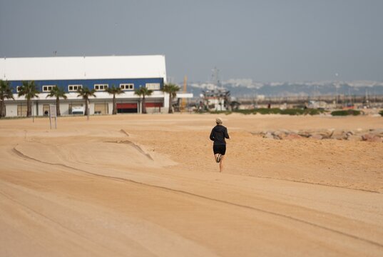Full length of athletic woman running along seashre. Side view of young sportswoman in white top and sporty shorts jogging on sandy beach. Fit lady represents healthy lifestyle.