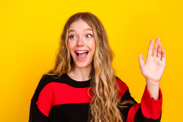 Cheerful blonde woman waving and smiling wearing a trendy striped sweater against a vibrant yellow...