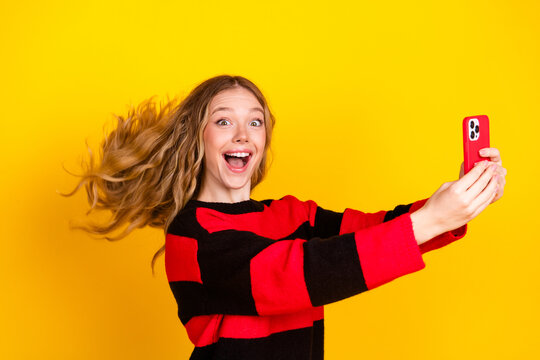 Cheerful young woman with blonde hair taking a selfie on her smartphone against a vibrant yellow background