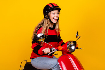 Cheerful young woman riding red scooter in casual sweater against bright yellow background,...