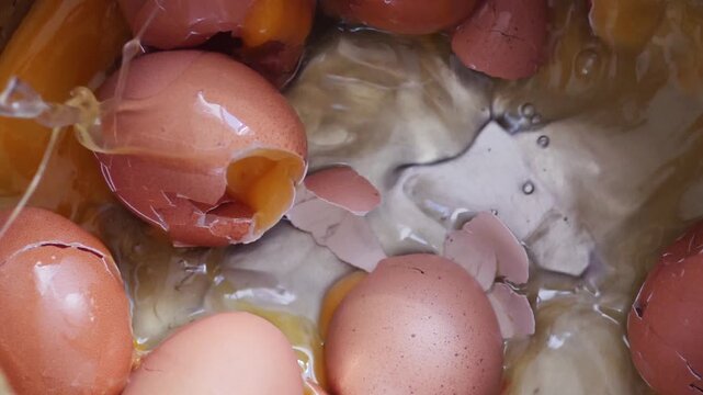 Multiple raw brown chicken eggs fall into a metal pot in a close-up overhead shot containing cracked shells. Upon impact, the shells break, causing fresh egg whites 