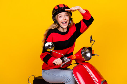 Cheerful young woman enjoying a ride on a red scooter against a bold yellow background in stylish casual knitwear
