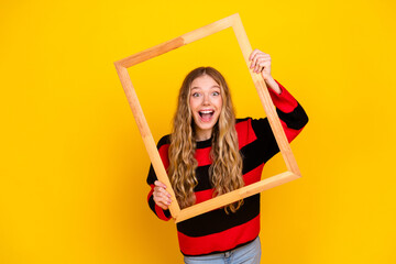 Cheerful young woman holding a wooden frame against a bright yellow background, expressing happiness