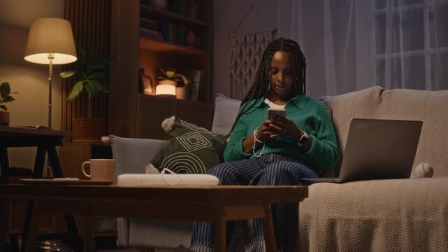 African American young adult woman sitting on sofa at night, using smartphone and laptop, then entering credit card details. Video is ideal for e-commerce, online banking, or digital burnout ads