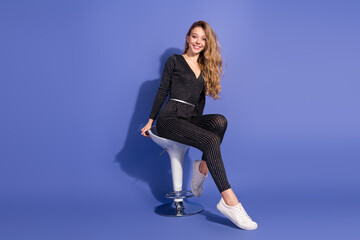 Young woman in striped jumpsuit sits on modern white bar stool against a blue studio background