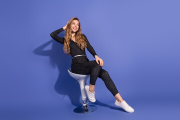 Young stylish woman sits on a modern white stool against a blue background wearing a striped...