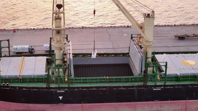 Cargo ship using deck cranes to load white bags from trucks at the port terminal. Maritime logistics, industrial shipping, and international freight transport operations.