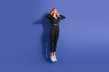 Young stylish woman in striped jumpsuit wearing pink headphones floats in mid air against a blue...