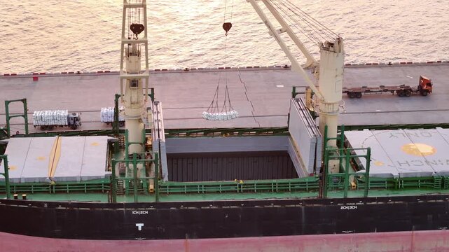 Cargo ship using deck cranes to load white bags from trucks at the port terminal. Maritime logistics, industrial shipping, and international freight transport operations.
