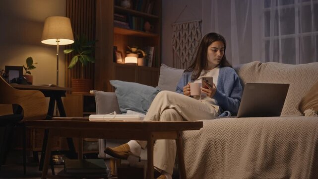 Young adult Caucasian woman sitting on sofa at night, holding mug and using smartphone before looking at laptop. Footage represents digital burnout and remote work fatigue concepts