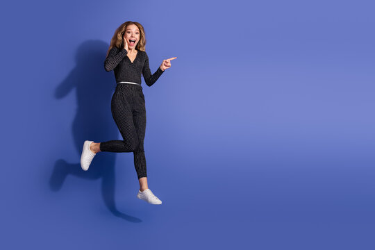 Joyful woman in striped jumpsuit jumps against blue background smiling and pointing for a playful fashion lifestyle image